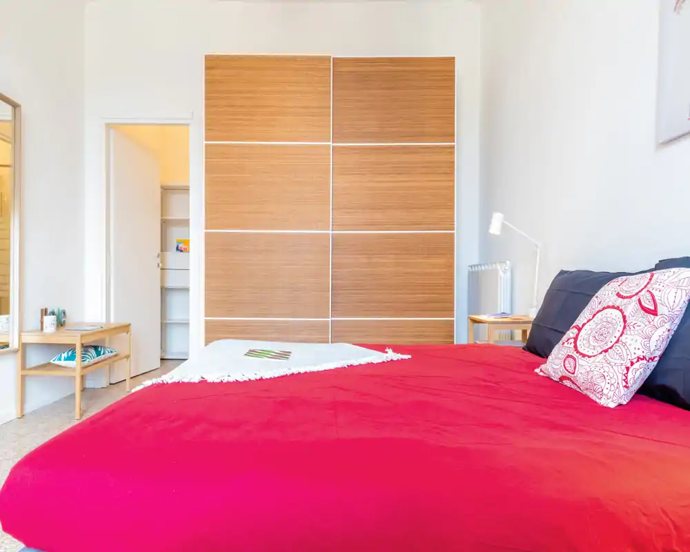Chambre lumineuse et moderne avec un grand lit rouge au premier plan, une armoire coulissante en bois et des meubles minimalistes pour une ambiance propre et chaleureuse.