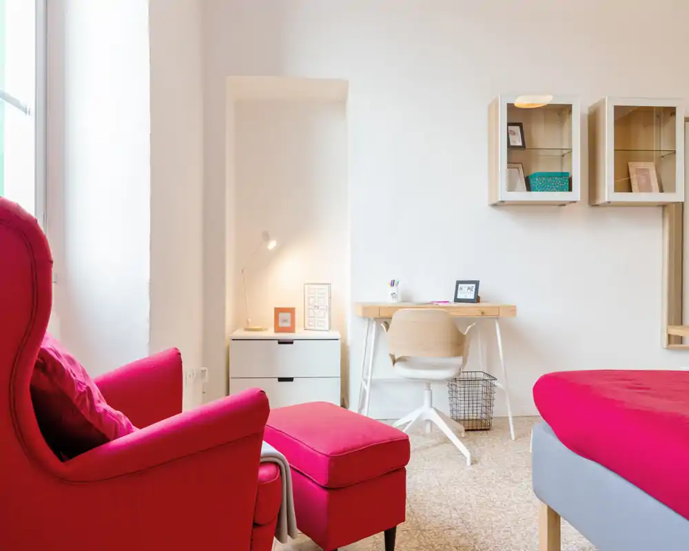 Coin chambre lumineux et moderne avec lit rouge visible et fauteuil assorti, petit bureau et étagères — idéal pour un séjour chaleureux.