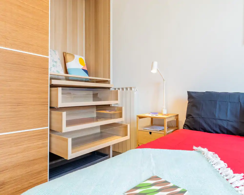 Coin chambre lumineux et moderne avec lit partiellement visible, table de chevet et tiroirs en bois ouverts ; linge propre et bois chaleureux pour une ambiance accueillante.