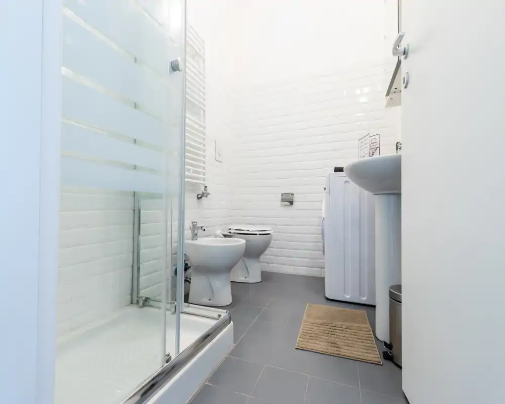 Salle de bains moderne et lumineuse avec cabine de douche en verre, bidet, WC et lavabo sur un sol carrelé gris ; murs en carreaux métro blancs et une machine à laver compacte pour un espace propre et fonctionnel.