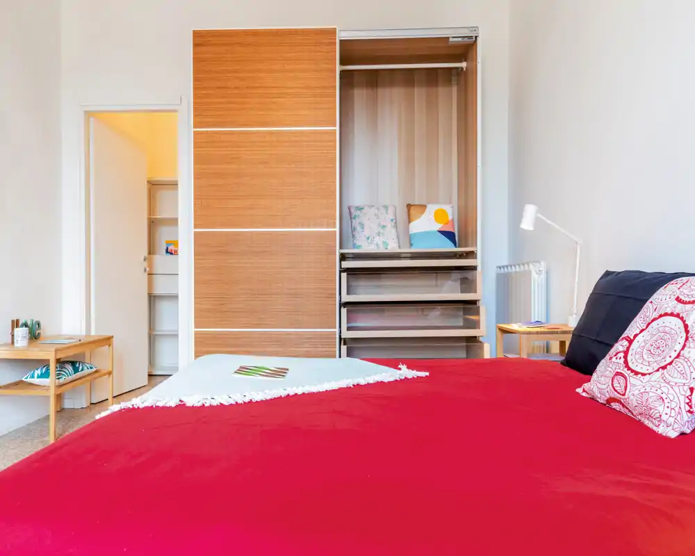 Chambre lumineuse et bien mise en scène avec un grand lit rouge au premier plan, tables de chevet et une armoire ouverte avec des coussins — photo idéale pour une annonce mettant en avant confort et rangement.
