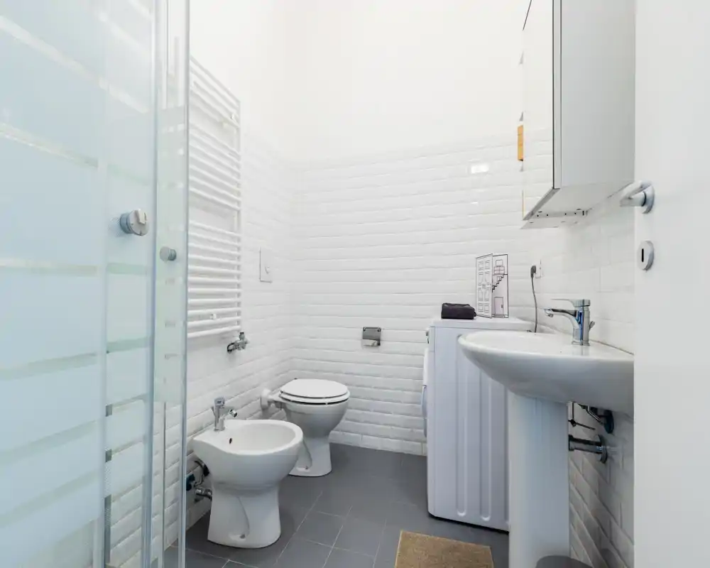 Salle de bains moderne et propre avec carrelage métro blanc, toilettes, bidet, lavabo sur colonne et douche à verre dépoli. Espace lumineux, minimaliste et soigné, adapté à une annonce locative.