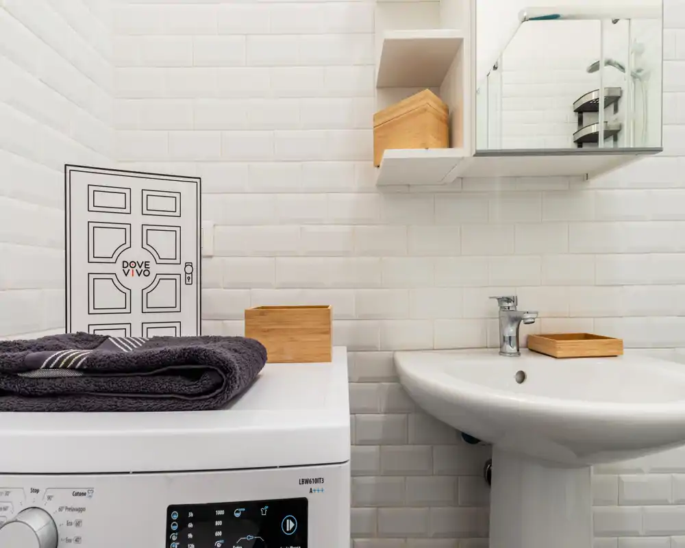 Coin de salle de bains propre et compact avec lavabo sur pied, armoire miroir et murs carrelés ; le dessus du lave-linge accueille des serviettes pliées et des boîtes en bois.