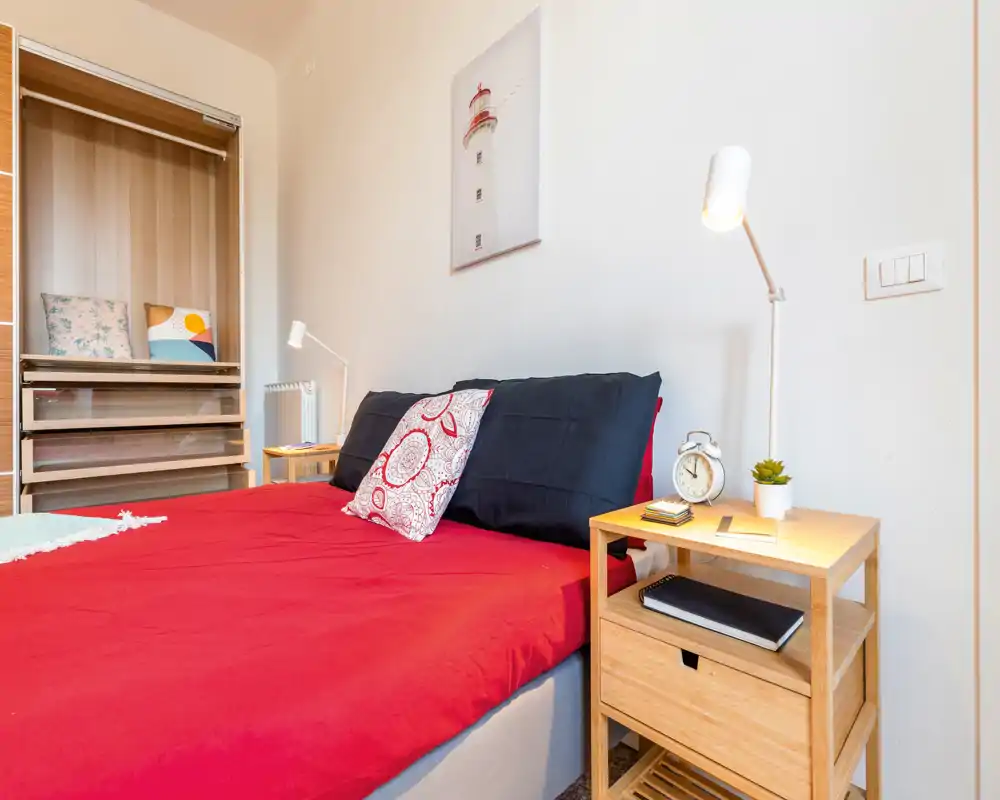 Chambre lumineuse et bien décorée avec housse de couette rouge, coussins décoratifs et table de chevet en bois avec lampe et réveil — idéale pour les photos d’annonce.
