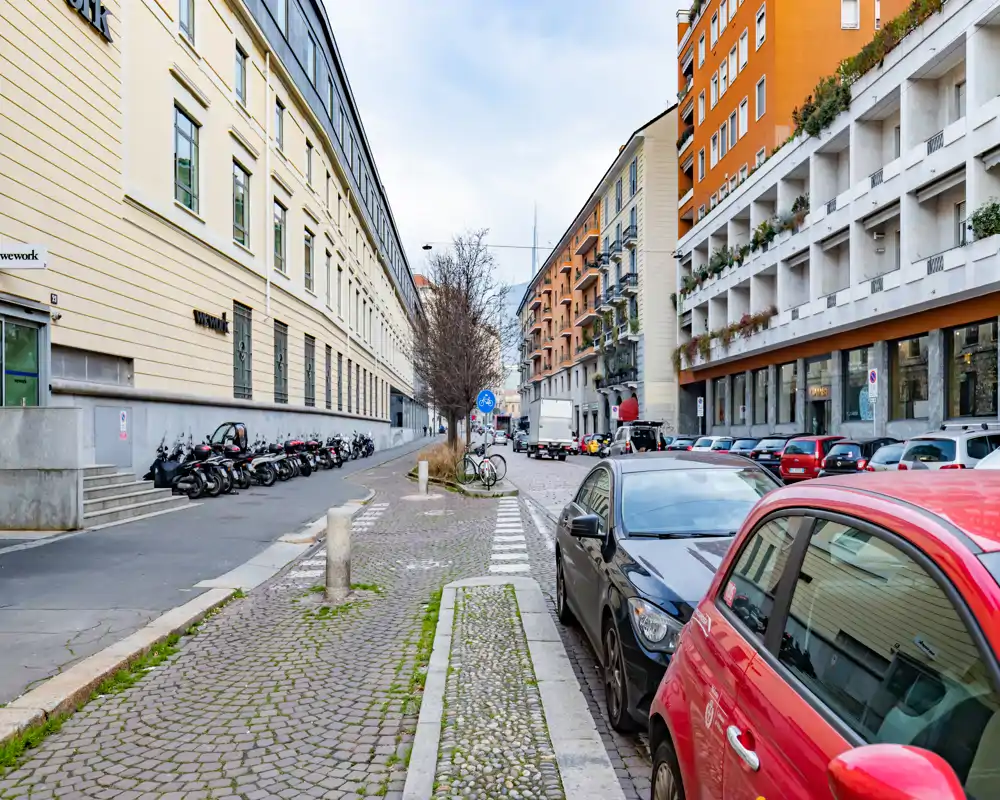 Vue de rue urbaine avec trottoir et voitures garées, immeubles résidentiels et commerciaux de taille moyenne, piste cyclable et scooters visibles.