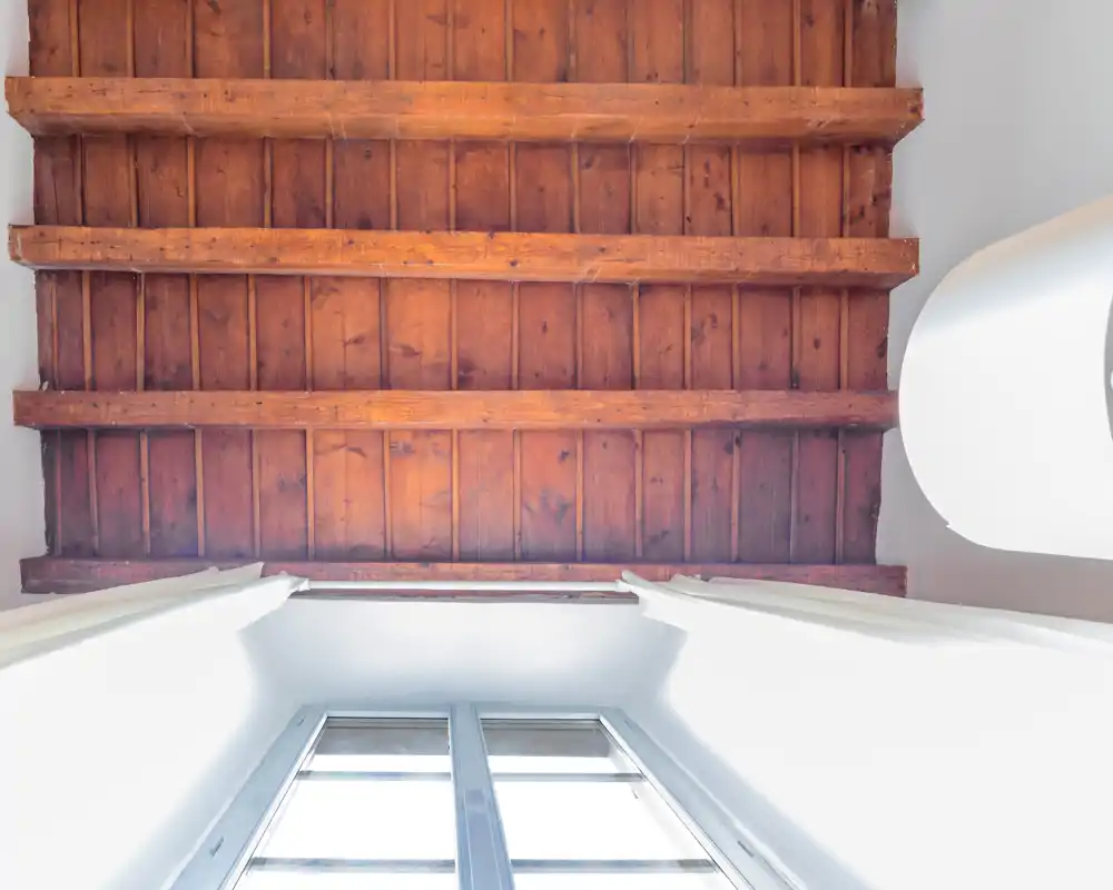 Vue vers le haut d'un plafond à poutres en bois au-dessus d'une fenêtre lumineuse ; détail architectural avec bois apparent et murs blancs.