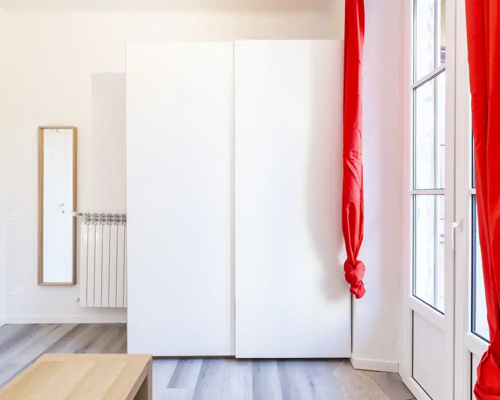 Angolo di camera minimal con un grande armadio bianco, specchio a figura intera, pavimento in legno e tende rosse vivaci vicino a una porta finestra che lascia entrare luce naturale.