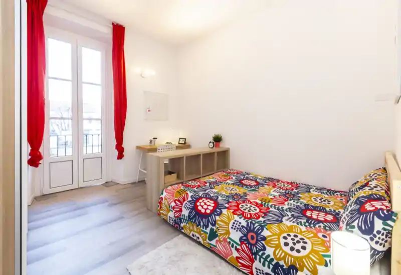 Camera da letto luminosa e colorata con letto ampio dal motivo floreale, luce naturale dalle porte-finestre, scrivania ordinata e dettagli decorativi accoglienti.