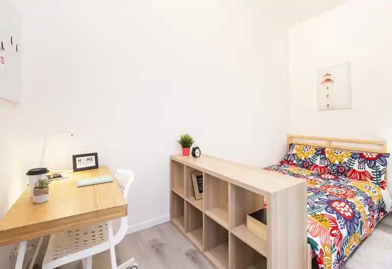 Camera da letto luminosa e compatta con un letto matrimoniale colorato, una scrivania in legno e scaffalatura divisoria — ideale per uno studente o una persona singola che cerca un ambiente allegro e ben illuminato.