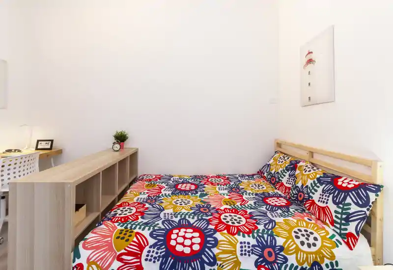 Camera da letto luminosa e compatta con una coperta floreale colorata, struttura del letto in legno semplice e mensole minimal in stile scandinavo per un ambiente riposante e ordinato.