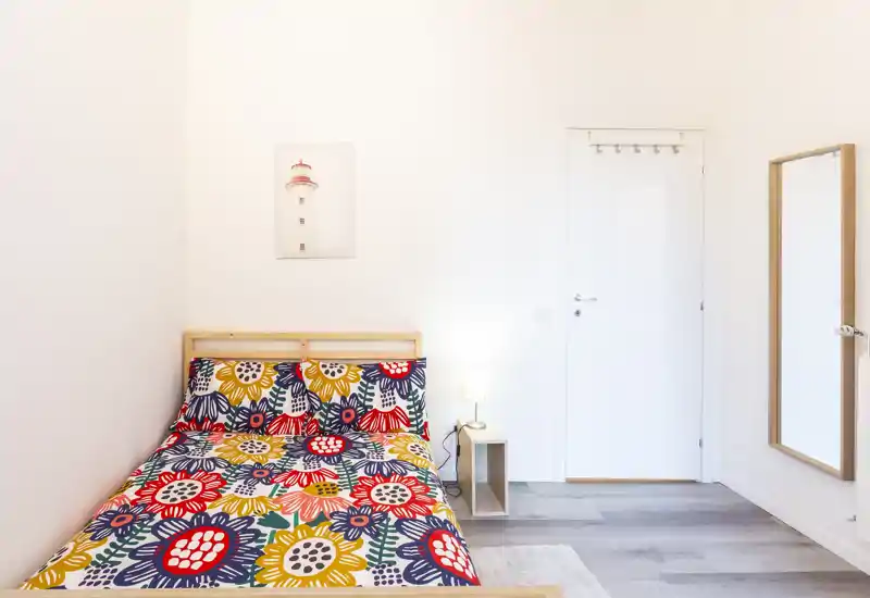 Camera da letto luminosa e moderna con un copriletto floreale colorato, una semplice struttura del letto in legno e una lampada da comodino. Pareti bianche pulite e uno specchio a figura intera rendono l'ambiente fresco e arioso.