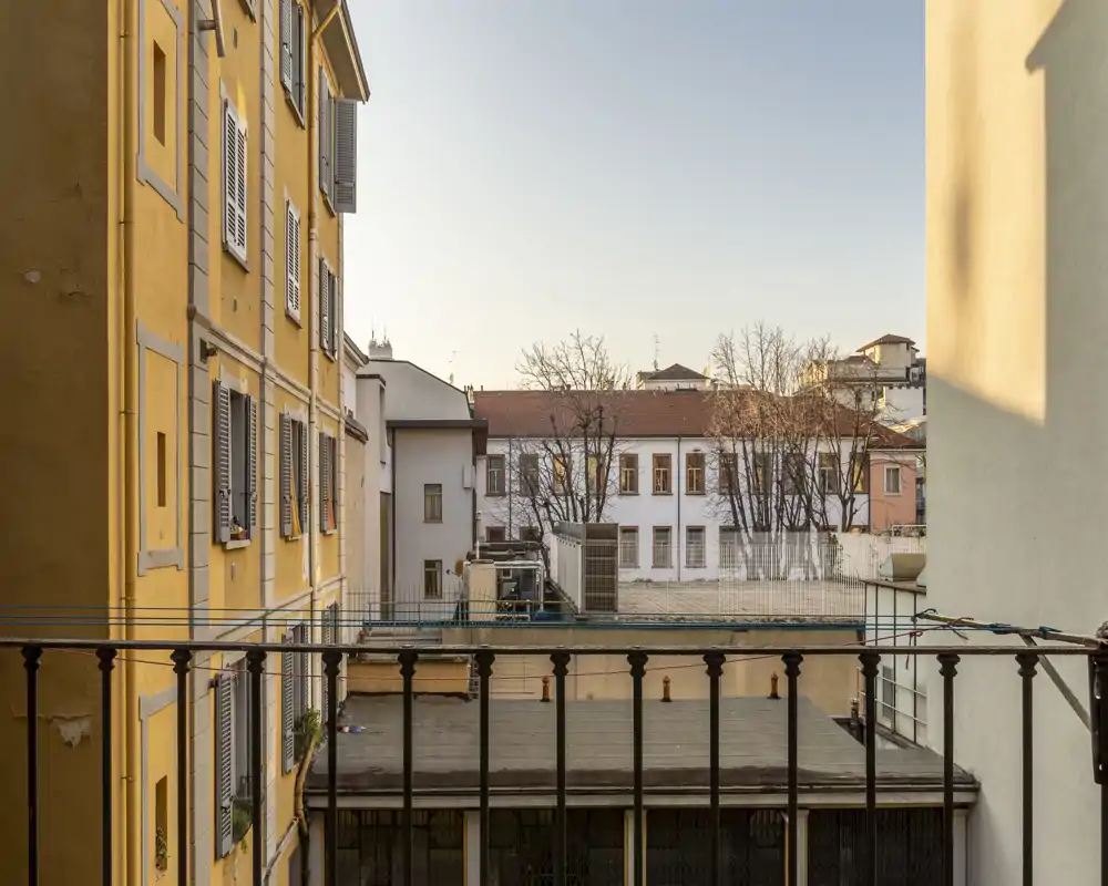 Vista dal balcone soleggiata su edifici limitrofi e cortile, con calda luce pomeridiana e ringhiera in ferro battuto in primo piano.