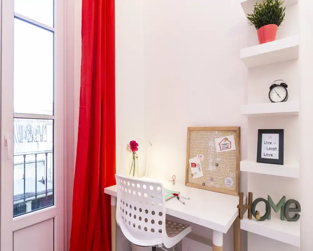 Angolo studio luminoso e ordinato con scrivania bianca, sedia moderna traforata, pannello in sughero e dettagli decorativi accanto a una finestra alta con tende rosse.