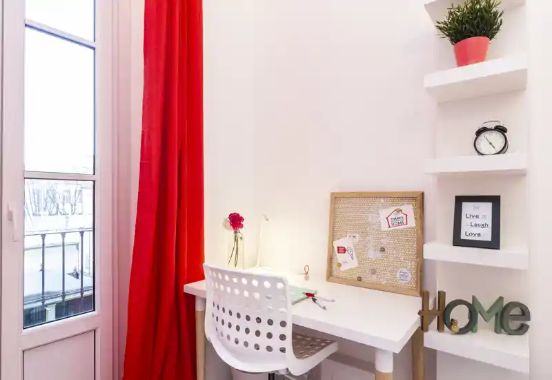 Coin bureau lumineux et soigné avec un bureau blanc, une chaise moderne ajourée, un tableau en liège et des accents décoratifs près d'une grande fenêtre aux rideaux rouges.