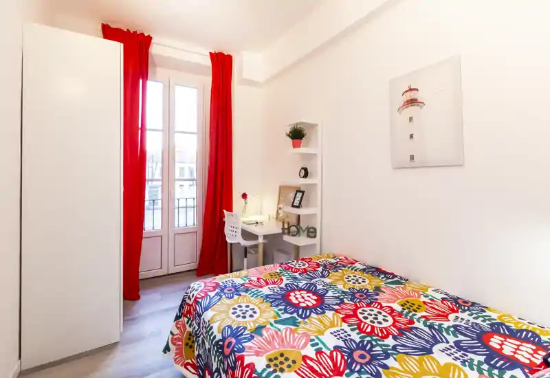 Camera da letto luminosa e compatta con copriletto floreale colorato, scrivania vicino alla finestra e tende rosse che rendono lo spazio vivido e ben illuminato, ideale per una persona.