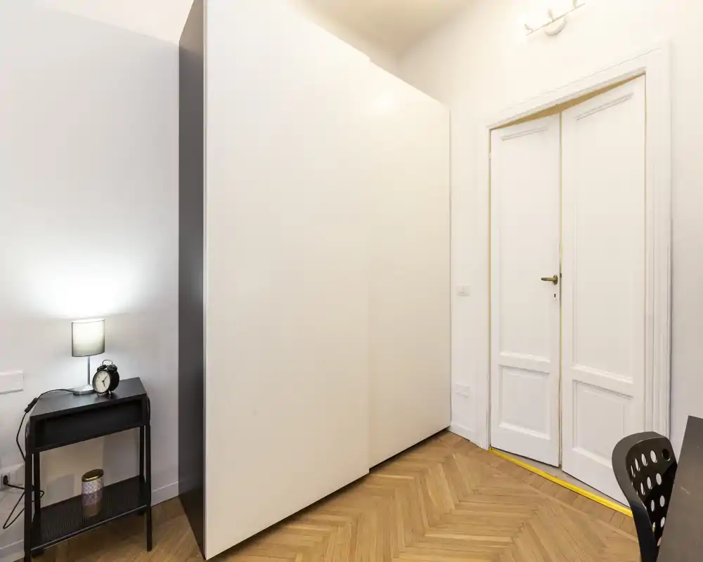 Coin de chambre lumineux et minimaliste avec une grande armoire blanche, une porte blanche et une petite table de chevet avec lampe et réveil.