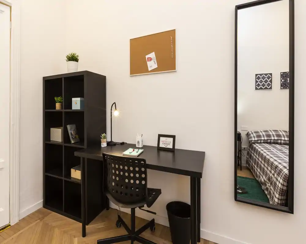 Coin chambre avec espace bureau : bureau noir et chaise, étagère et grand miroir mural reflétant un lit simple bien fait avec une couette à motifs.
