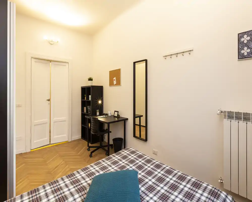 Chambre bien éclairée avec couvre-lit à motifs, coussin turquoise et un bureau compact avec étagères — espace confortable et fonctionnel, parfait pour une personne.
