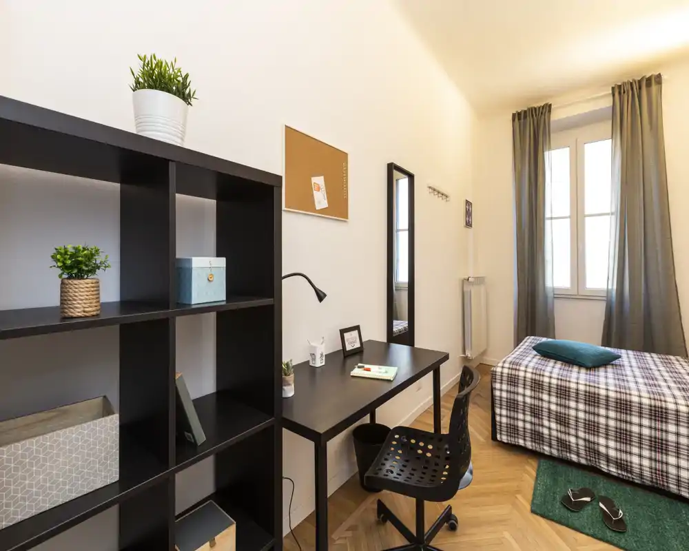 Chambre lumineuse et soignée avec un lit simple, un bureau et des étagères — idéale pour un étudiant ou une personne seule. La lumière naturelle et le parquet chaleureux apportent une atmosphère accueillante.