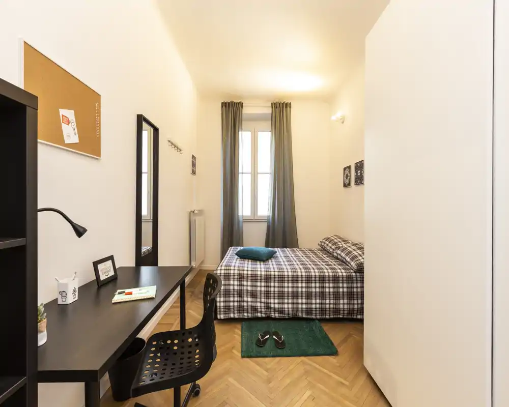 Chambre simple lumineuse et bien aménagée avec lit à carreaux, bureau et lumière naturelle douce — idéale pour étudiant ou professionnel.