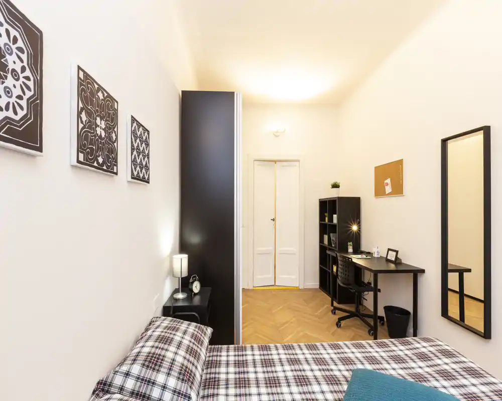 Chambre lumineuse et moderne avec couvre-lit à motifs, lampe de chevet et un espace de travail avec bureau et étagères — idéale pour se reposer et travailler.
