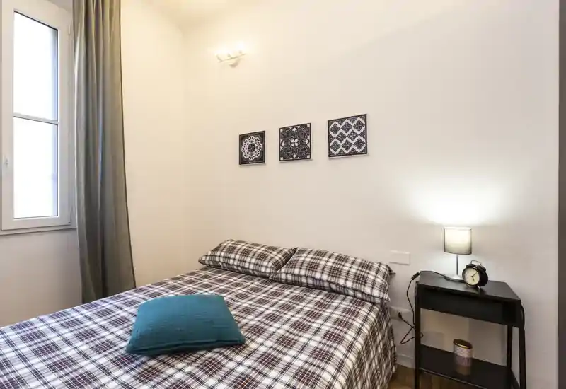 Chambre confortable et lumineuse avec un lit double en couvertures à carreaux, coussin bleu sarcelle, table de chevet avec lampe et réveil, et décoration murale simple.