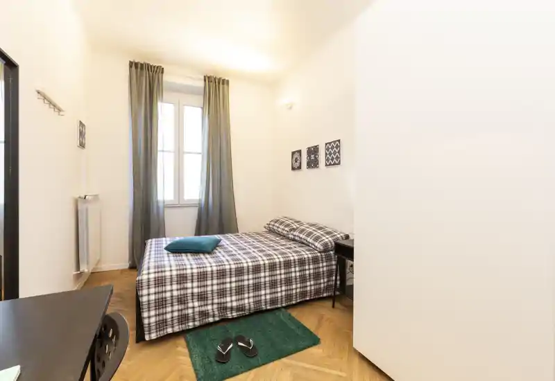 Chambre lumineuse et soignée avec un lit double à couvre-lit à carreaux, une table de chevet et une fenêtre avec rideaux apportant de la lumière naturelle.