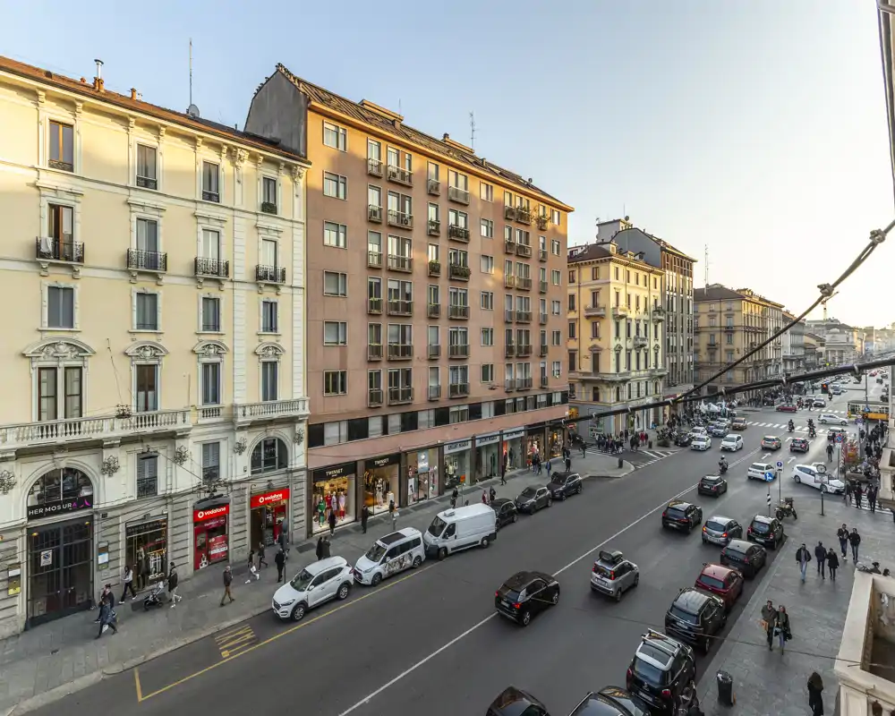 Vista amplia de la calle que muestra una avenida urbana concurrida con coches aparcados, peatones y edificios de varias plantas bañados por una cálida luz de media tarde.