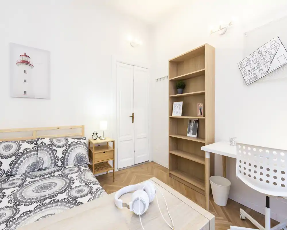 Dormitorio luminoso y bien decorado con cama doble, mesita de noche y una pequeña zona de trabajo. Suelo de madera y paredes claras crean un ambiente acogedor y fresco.