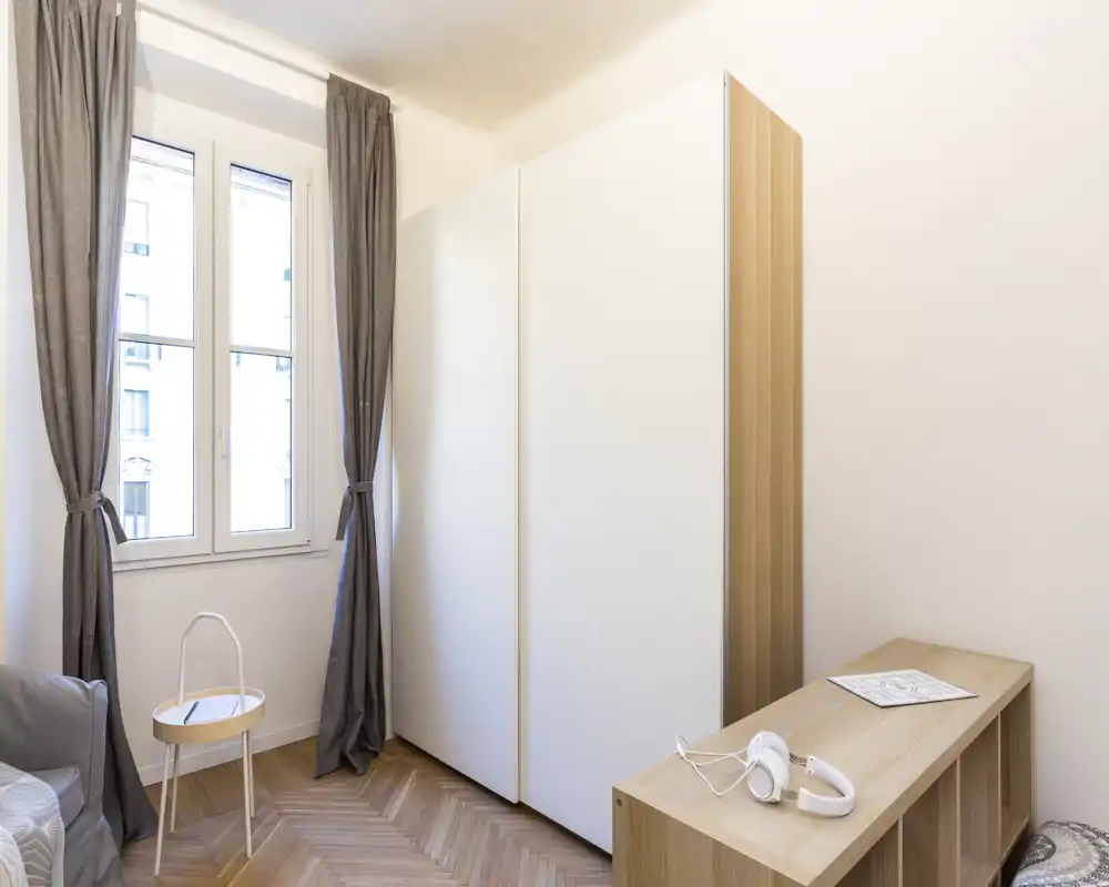 Rincón de dormitorio luminoso con un gran armario blanco, luz natural desde la ventana y muebles de madera sencillos que crean un estilo limpio y minimalista.