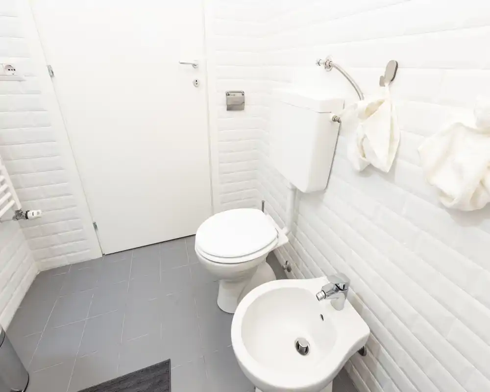 Bagno pulito e luminoso con piastrelle bianche effetto subway, WC e bidet e piastrelle grigie a pavimento. Lo spazio è ben illuminato e minimale, perfetto per un annuncio moderno.