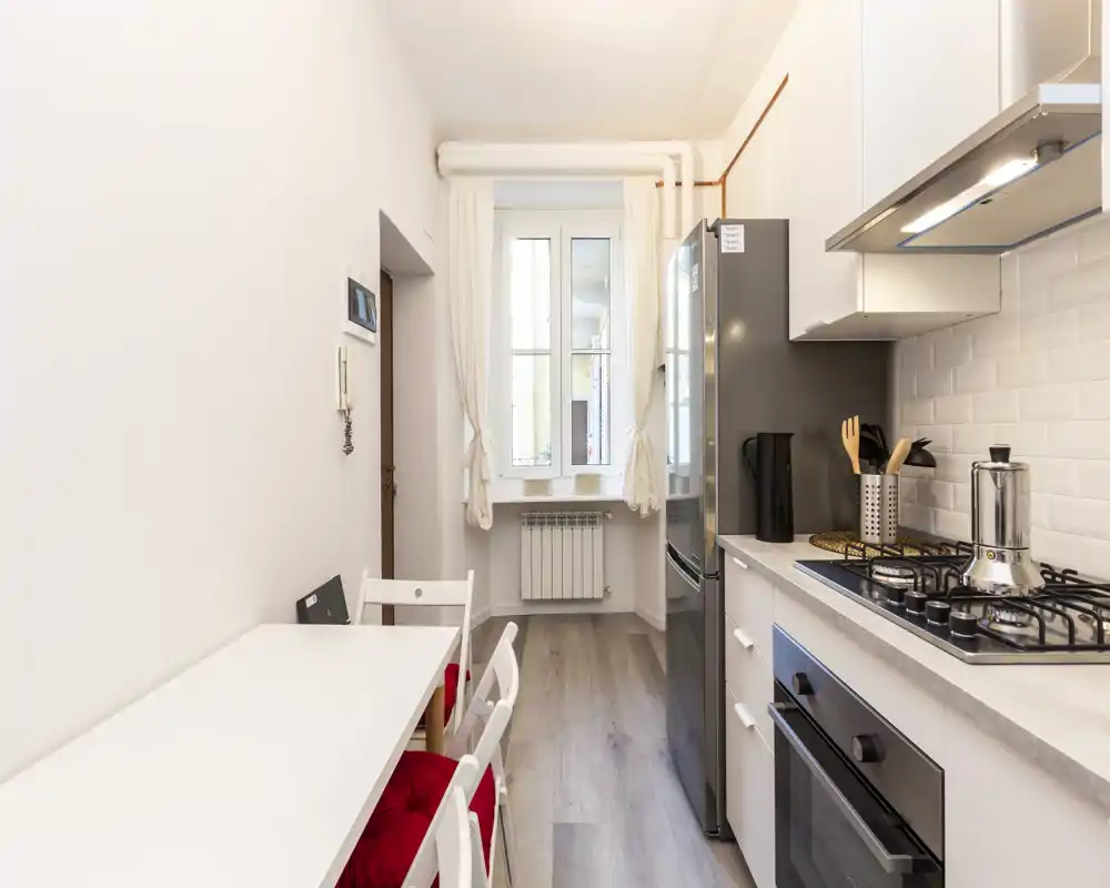 Cucina moderna e luminosa, stretta, con piano cottura a gas, forno e frigorifero; piccolo tavolo con sedie e cuscini rossi vicino alla finestra che offre luce naturale.
