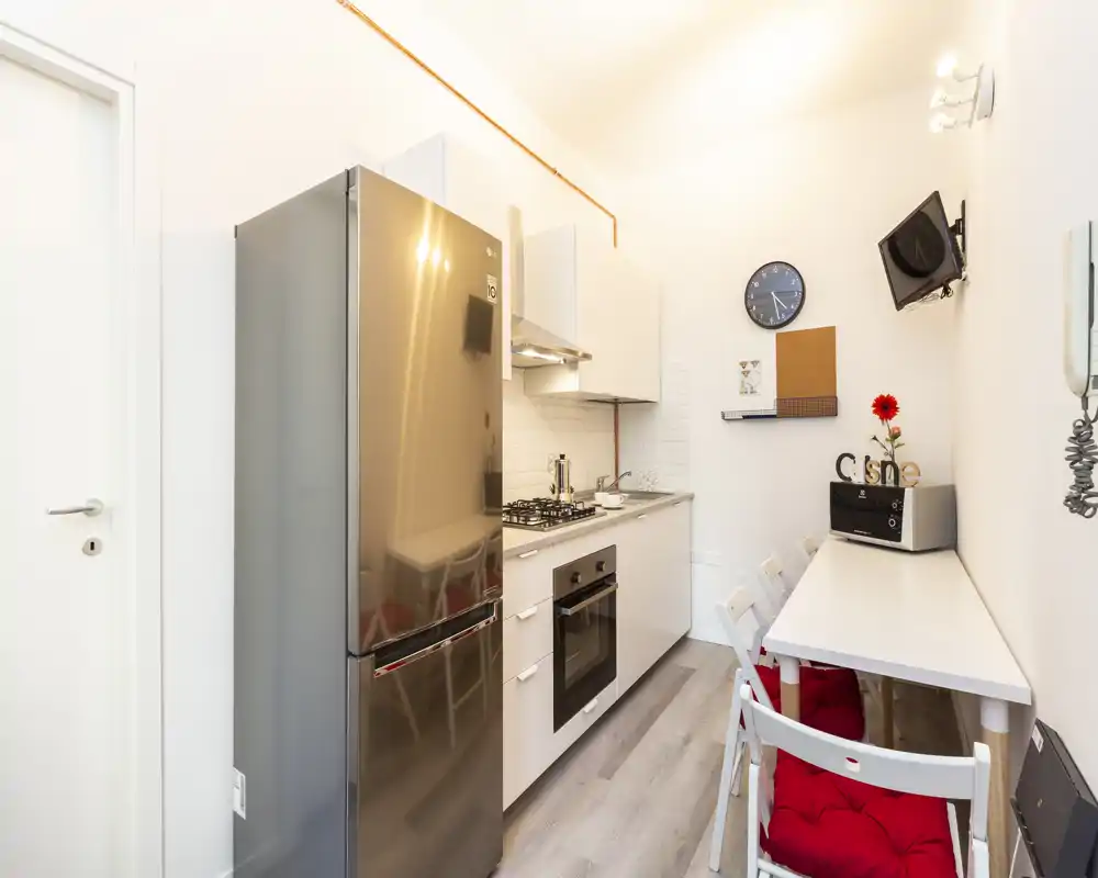 Cucina moderna e luminosa a corridoio con frigorifero in acciaio, forno incassato e piccolo bancone pranzo con cuscini rossi — compatta e organizzata per soggiorni brevi.