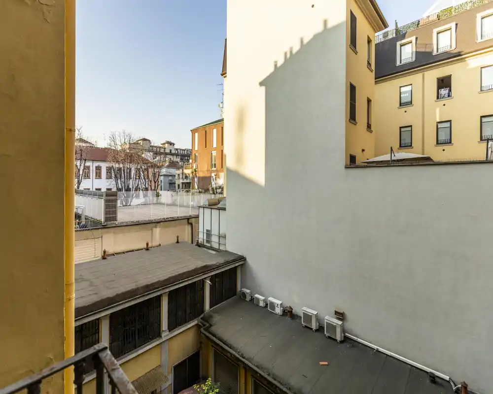 Vista su un cortile interno e le facciate degli edifici adiacenti vista da una finestra o un piccolo balcone; immagine in luce diurna che mostra tetti, unità di condizionamento e appartamenti vicini.