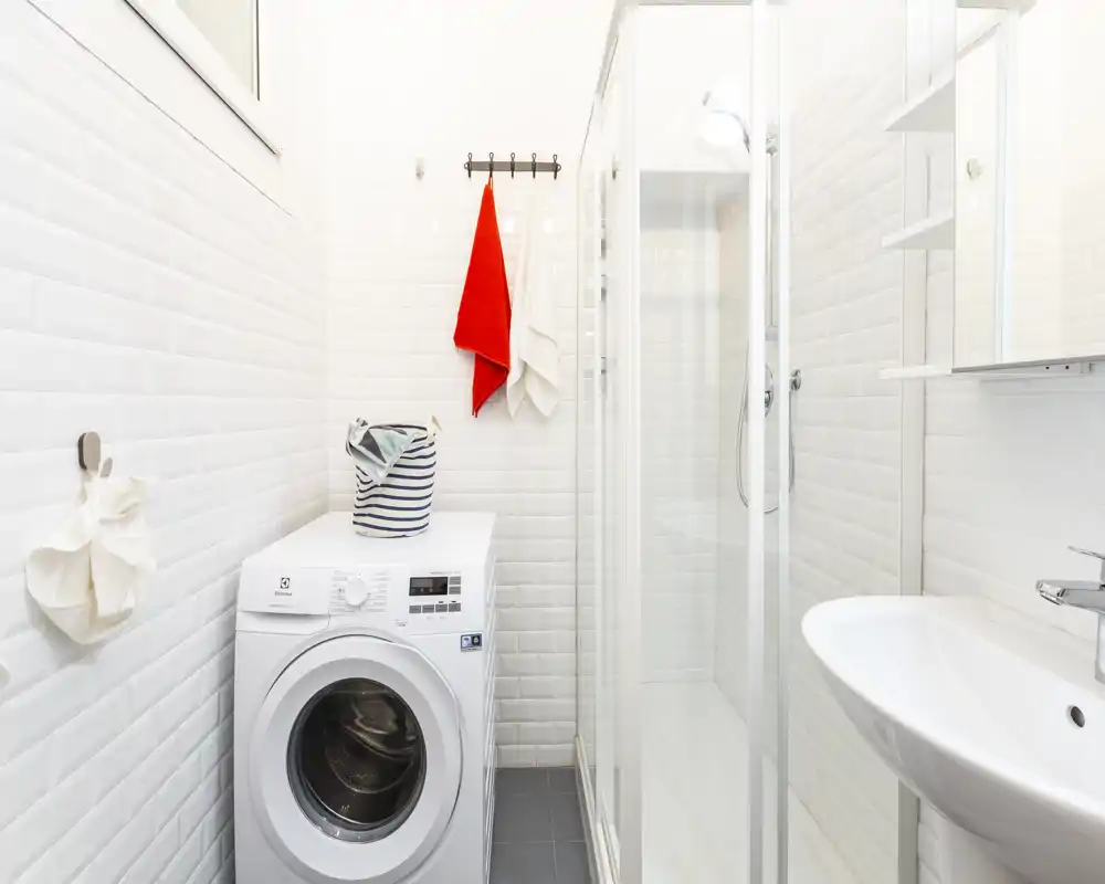 Bagno moderno e compatto con lavatrice a carica frontale, doccia con porta in vetro e lavabo a parete; piastrelle bianche e illuminazione intensa per un ambiente pratico e luminoso.