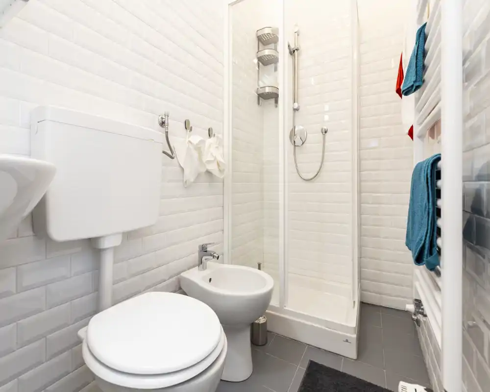 Bagno compatto e moderno con cabina doccia, WC, bidet e scalda-asciugamani; piastrelle bianche e pavimento grigio donano un aspetto pulito e fresco.