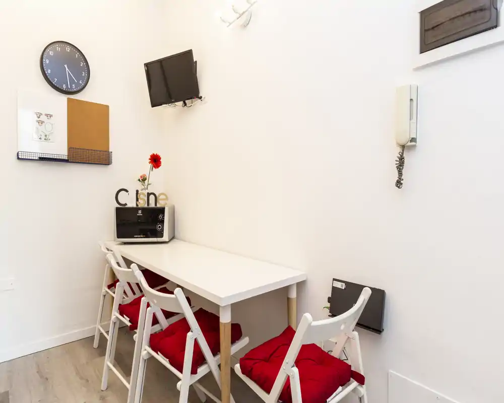 Angolo cucina compatto con tavolo bianco stretto, quattro sedie pieghevoli con cuscini rossi, microonde e TV a parete — luminoso e ordinato, ideale per pasti veloci o cucina leggera.