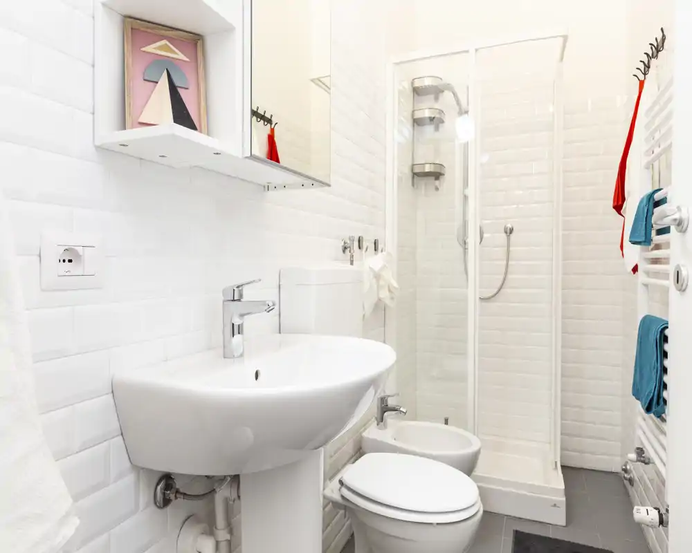 Bagno luminoso e moderno con lavabo a colonna, WC, bidet e doccia con box in vetro; piastrelle bianche effetto metro e termoarredo per un aspetto pulito e contemporaneo.