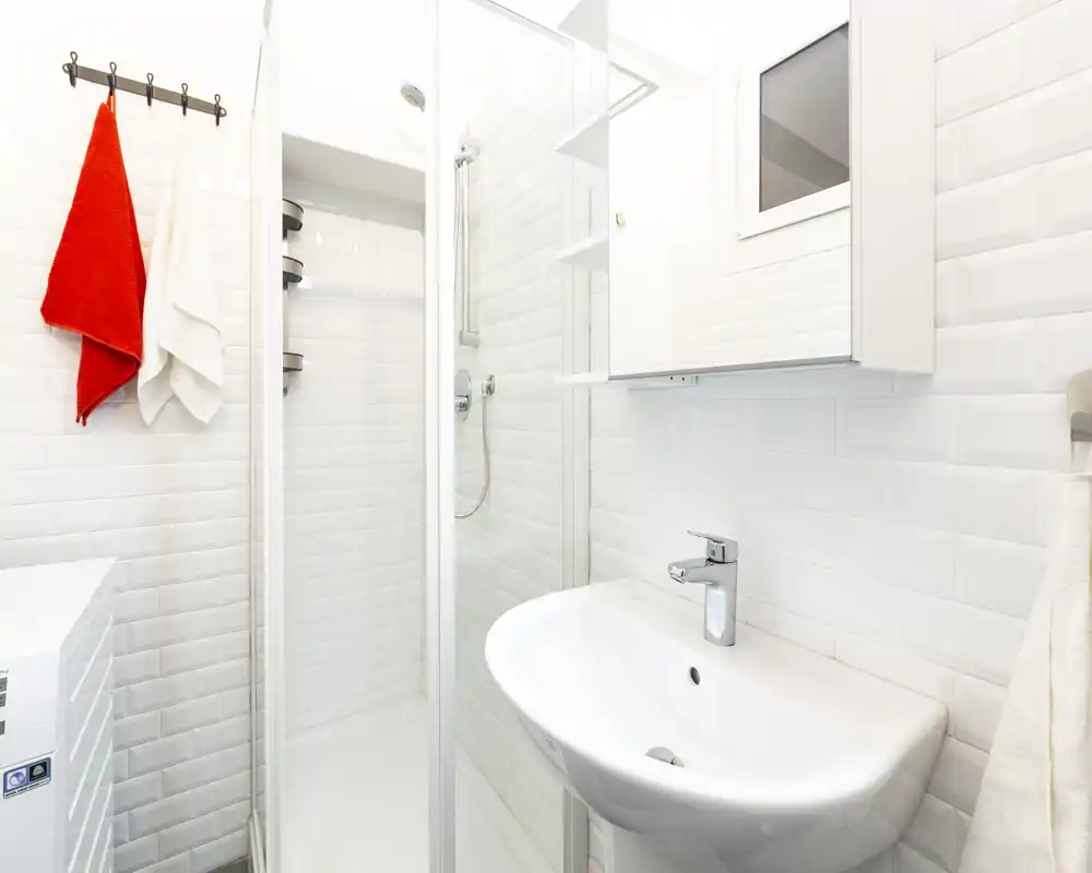 Bagno moderno e compatto con piastrelle bianche a muro, doccia angolare con porta in vetro e lavabo bianco: ambiente luminoso e pulito, adatto a affitti brevi.