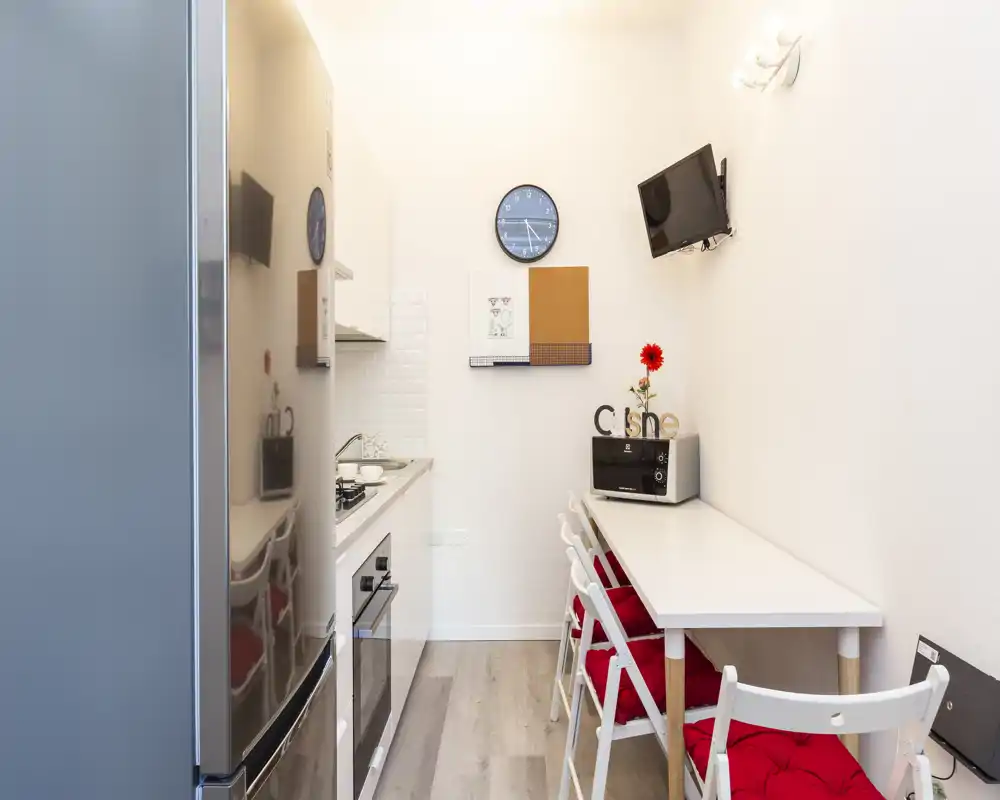 Cucina moderna compatta con forno incassato, piano cottura a gas e frigorifero; bancone stretto con sgabelli e cuscini rossi, pareti bianche luminose e dettagli decorativi.