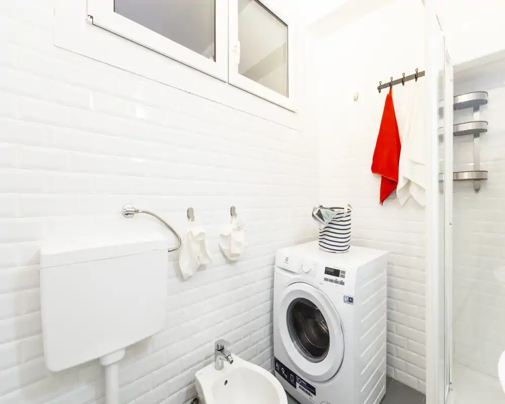 Bagno compatto e luminoso con piastrelle bianche, bidet, lavatrice e cabina doccia — pulito e funzionale.
