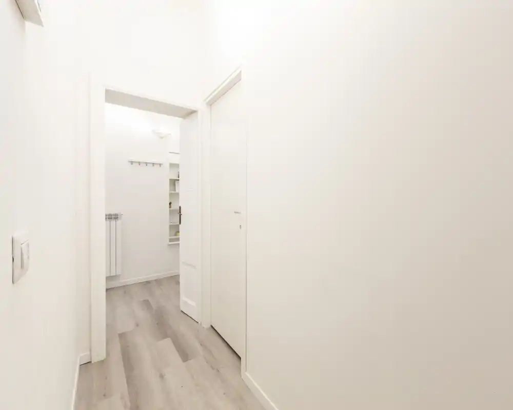 Ingresso/corridoio luminoso e minimale con pavimento chiaro in legno, pareti bianche e un appendiabiti visibile nell'alcova adiacente.