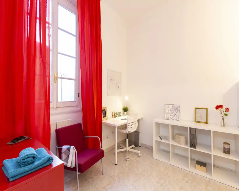 Angolo luminoso di camera/studio con scrivania, scaffalatura e una poltrona rossa vicino a una finestra con tende rosse vivaci.