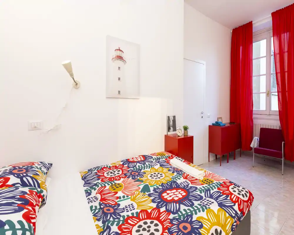 Camera da letto luminosa e vivace con un grande letto a fantasia floreale, tende rosse e arredi minimal. La stanza è ben illuminata da una finestra alta e arredata in stile moderno e allegro.