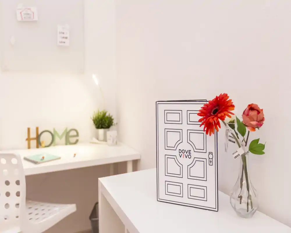 Angolo home office luminoso e minimal all'interno della zona giorno: scrivania bianca, insegna decorativa 'Home', lampada da scrivania e fiori freschi su un tavolino per un'atmosfera accogliente e moderna.