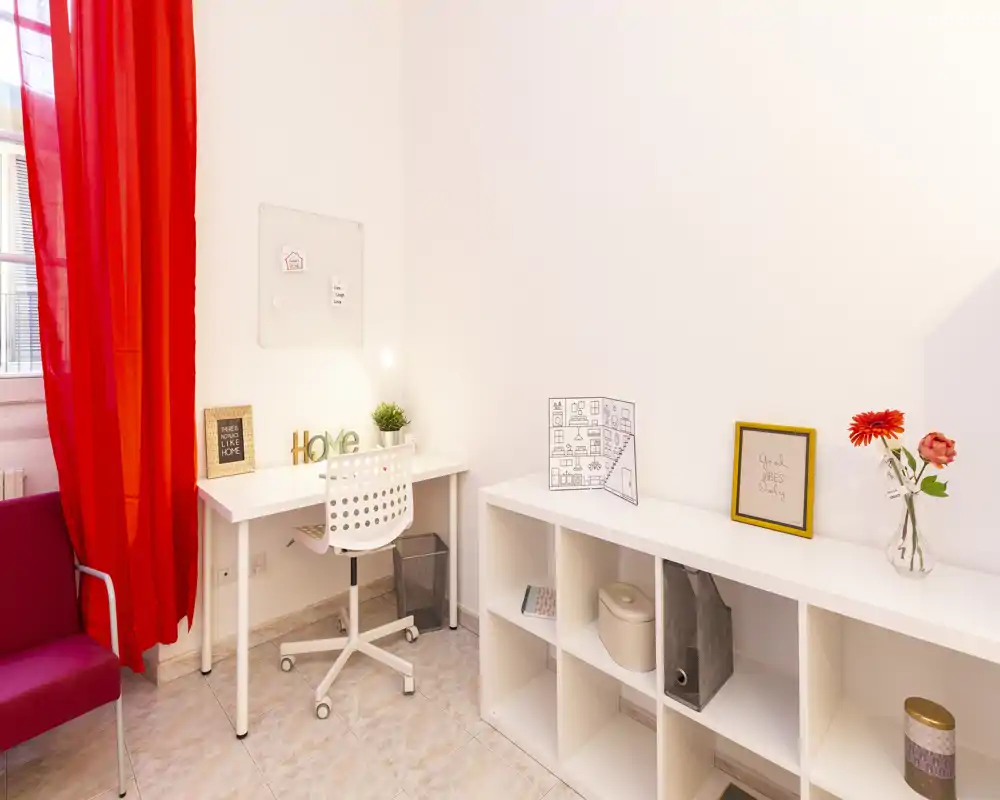 Spazio di lavoro domestico luminoso e moderno con scrivania bianca, sedia girevole e scaffalatura aperta; tende rosse e dettagli decorativi creano un'area accogliente e ideale per lo smart working.