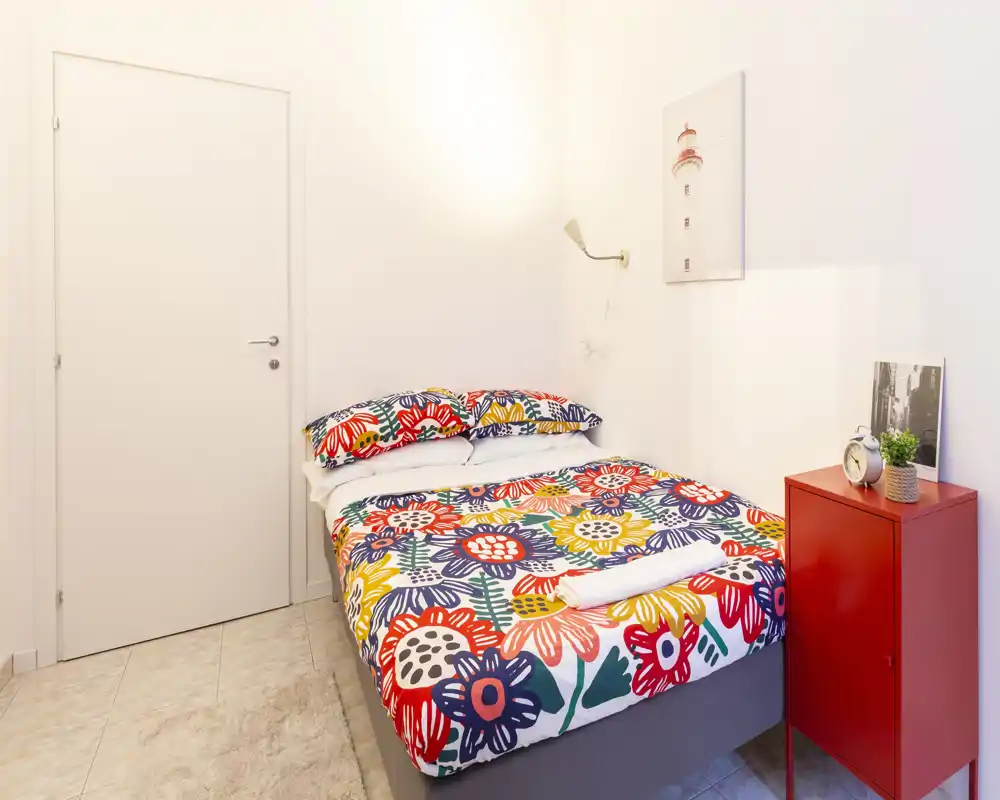 Camera da letto luminosa e accogliente con copriletto floreale colorato, comodino e lampada da parete — ideale per soggiorni singoli o ospiti.