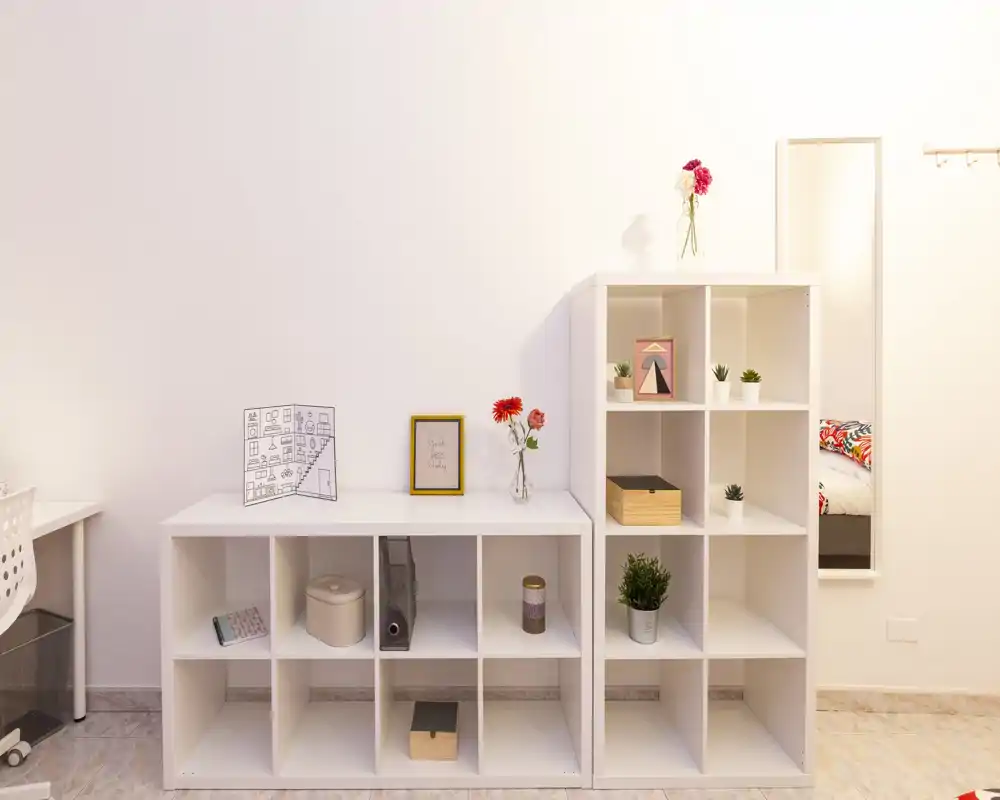 Angolo camera luminoso e minimale con scaffalatura modulare bianca, piccole piante decorative e un accenno di letto visibile solo nello specchio a parete. Stile pulito e moderno, adatto a un monolocale o camera singola.
