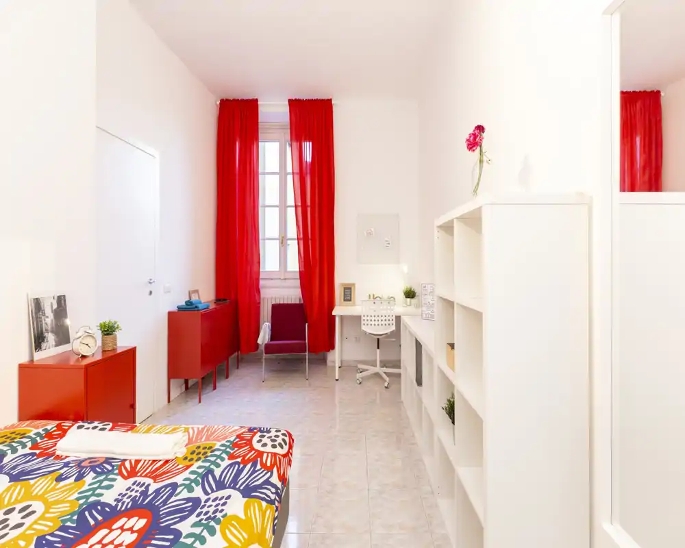 Camera da letto luminosa e colorata con copriletto a fantasia, tende rosse e una postazione di lavoro funzionale. La stanza è ordinata, ariosa e dotata di scaffalature e luce naturale dalla finestra.