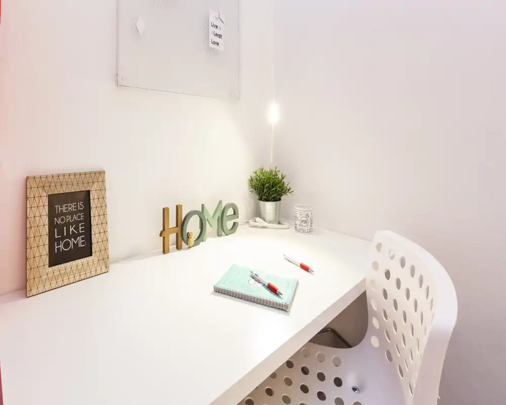 Angolo studio luminoso e minimal in camera con scrivania bianca, decorazione "HOME", pianta e quaderno — perfetto per uno spazio di lavoro tranquillo.